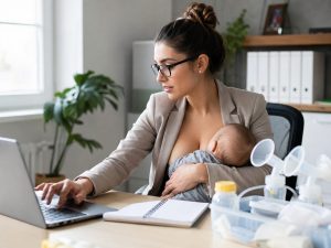 Madre trabajando con el portátil mientras amamanta a su bebé, con un sacaleches y biberones sobre la mesa